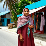 **A woman wearing a modest, stylish outfit including a shawl, standing on a Maldivian local island street. Background should feature local shops and people in traditional clothing, conveying a sense of respect for local culture.**